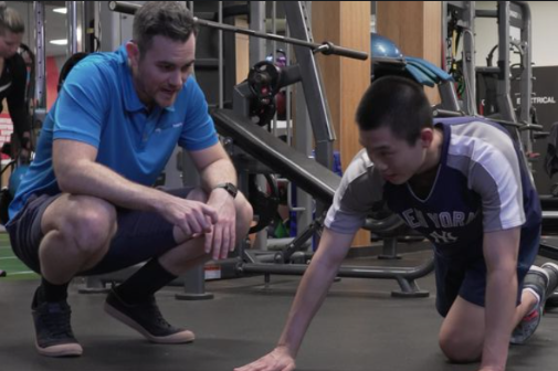 A fitness trainer in a blue polo shirt crouches down beside Liam on the gym floor, who is positioned on all fours. The trainer is engaged in a supportive conversation, pointing towards the ground as they work on exercise techniques at My Health Team in Redcliffe. 